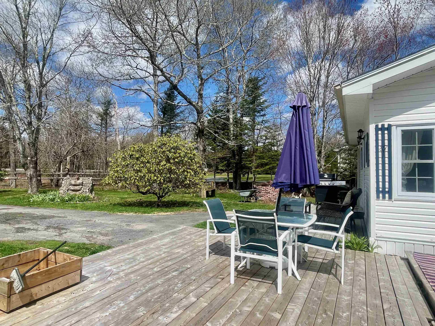 Outdoor patio with white table and chairs, a closed purple umbrella, and a wooden planter on a weathered deck. Trees and blue sky in the background.