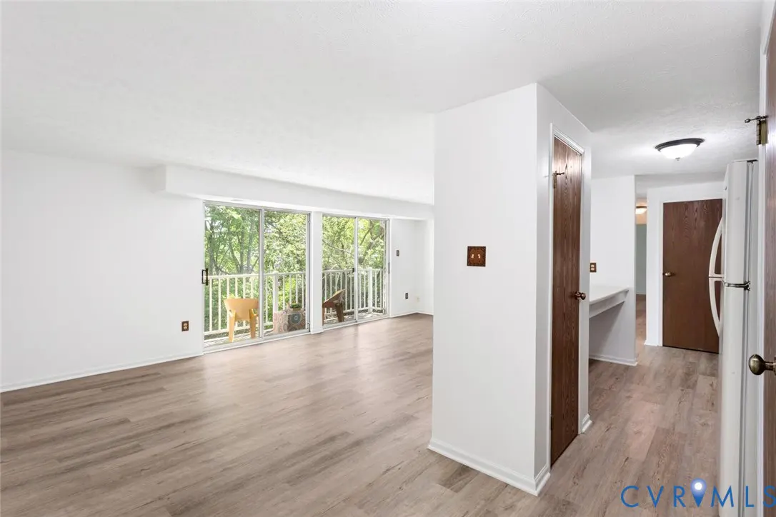 Bright, open living room with wood-look floors, white walls, and sliding glass doors to a balcony with green trees in the background.