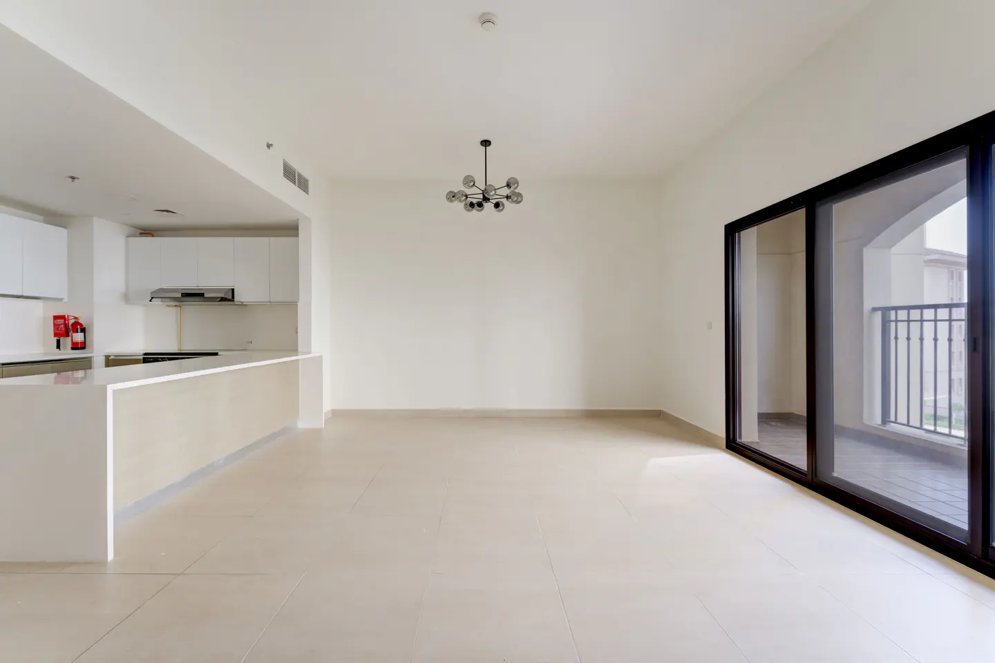 Bright, empty apartment with white walls, tile floor, and open kitchen. A black-framed sliding glass door leads to a balcony.