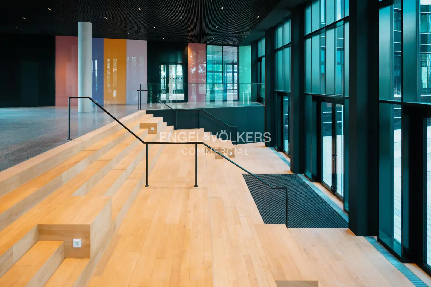 Interior view of a modern commercial space with light wood steps, black railings, and large teal-tinted windows. Colorful panels adorn the back wall.