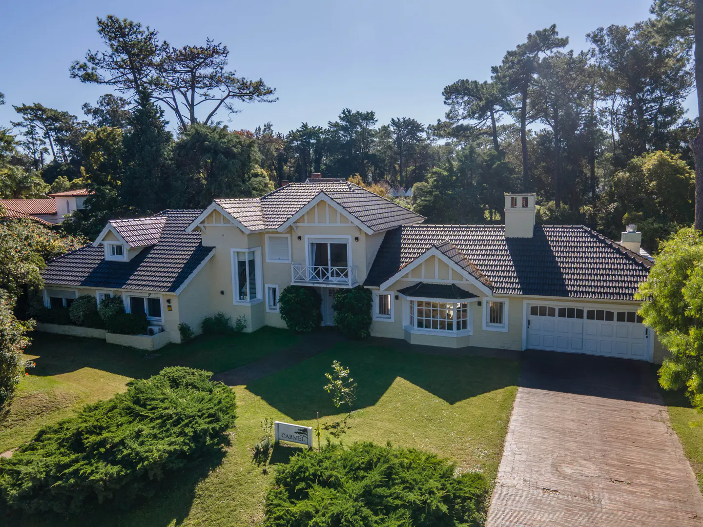Aerial view of a large, light yellow house with a dark tiled roof, surrounded by green trees and lawn. A brick driveway leads to a two-car garage.