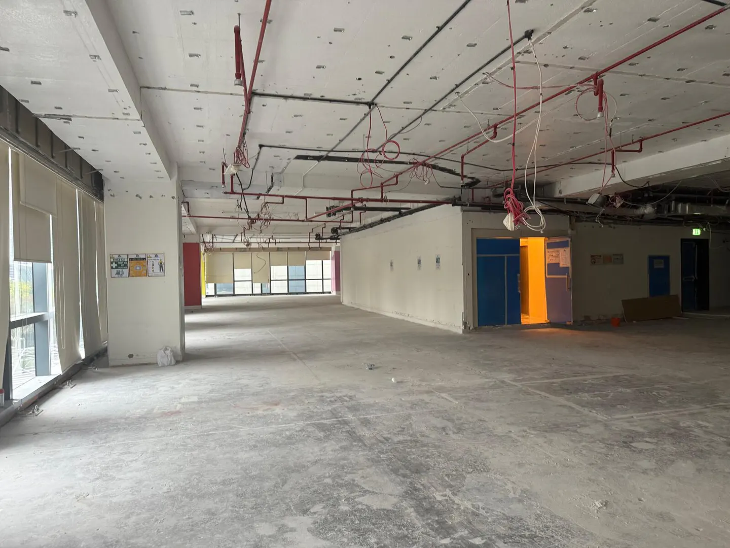 Unfinished office space with concrete floors, exposed ceiling pipes, and large windows with closed blinds.