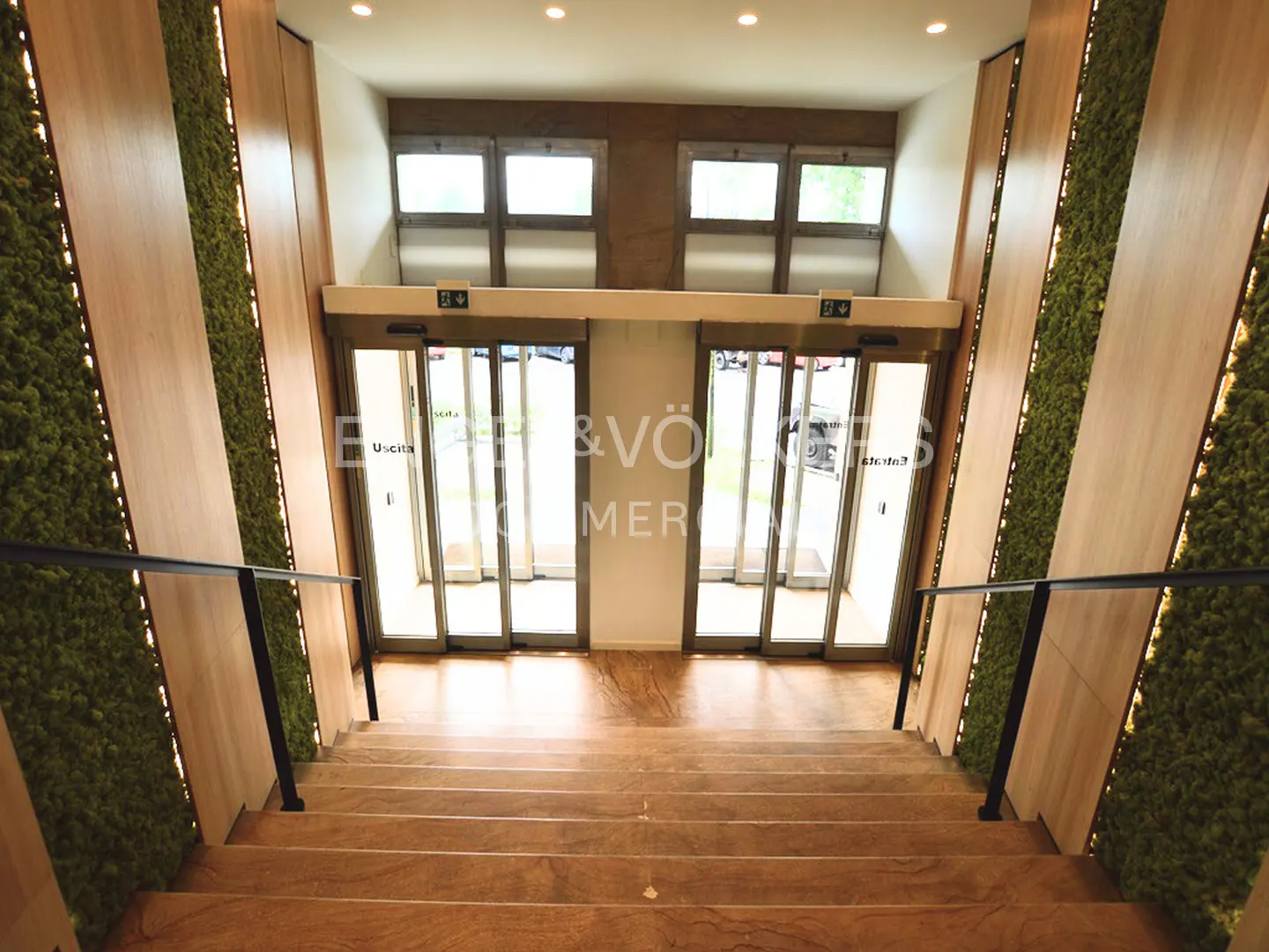 Stairway leading to glass doors with "Uscita" signs, flanked by moss walls and wood panels. Black handrails guide the way.