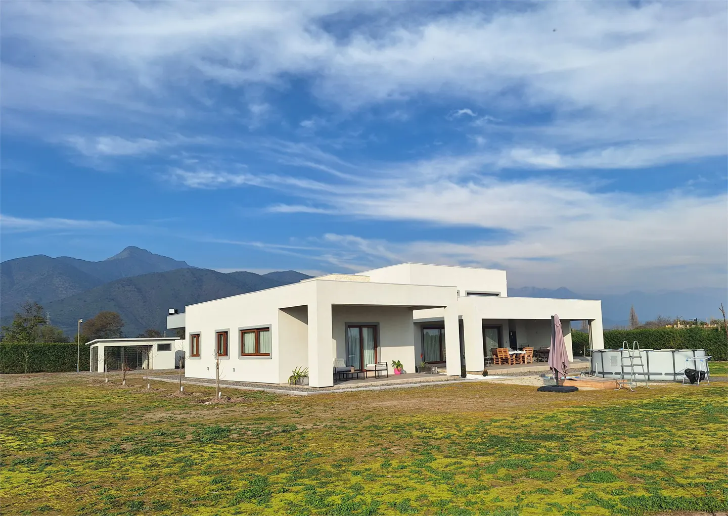Modern white house with brown window frames, a pool, and mountain views under a blue sky.