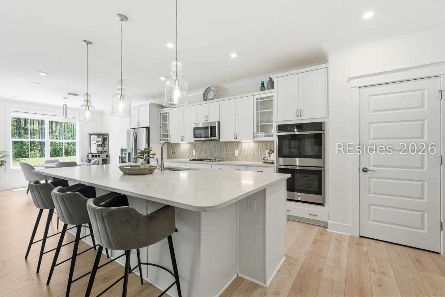 Bright, modern kitchen with white cabinets, stainless steel appliances, and a large island with gray bar stools. Hardwood floors and pendant lights.