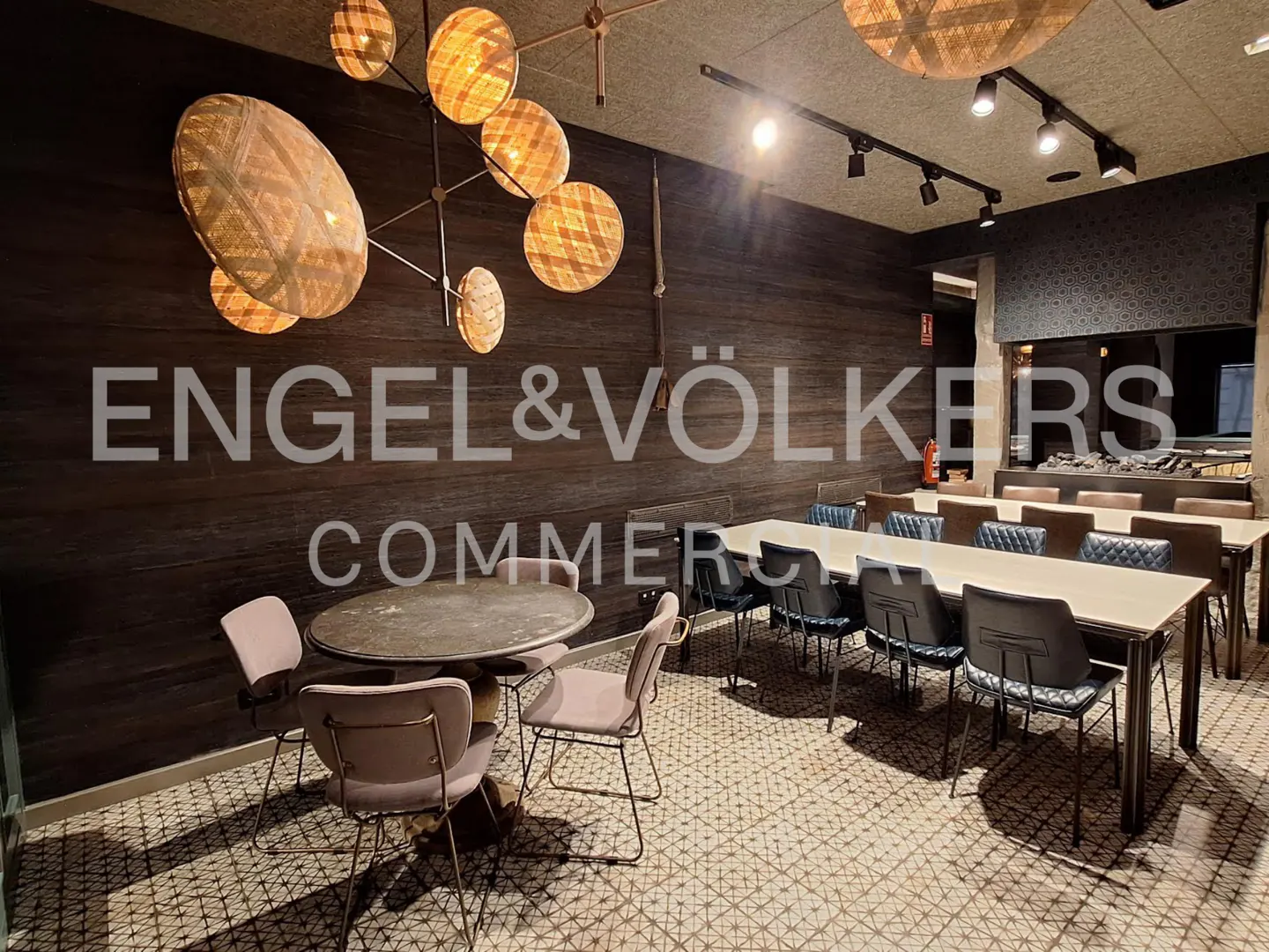 A commercial space with tables, chairs, and modern lighting fixtures. The walls are dark wood, and the floor has a geometric pattern.