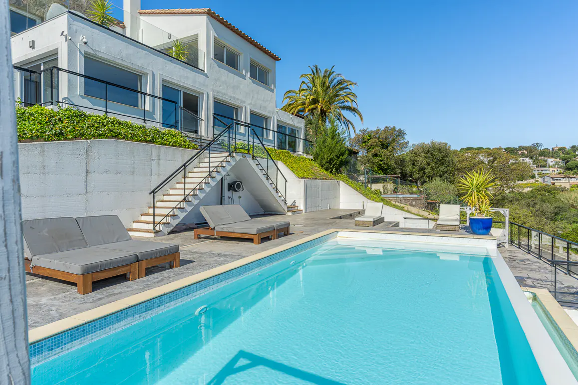A modern white house with a blue-tiled pool and gray lounge chairs on a sunny day. Stairs lead up to the house, surrounded by green foliage.