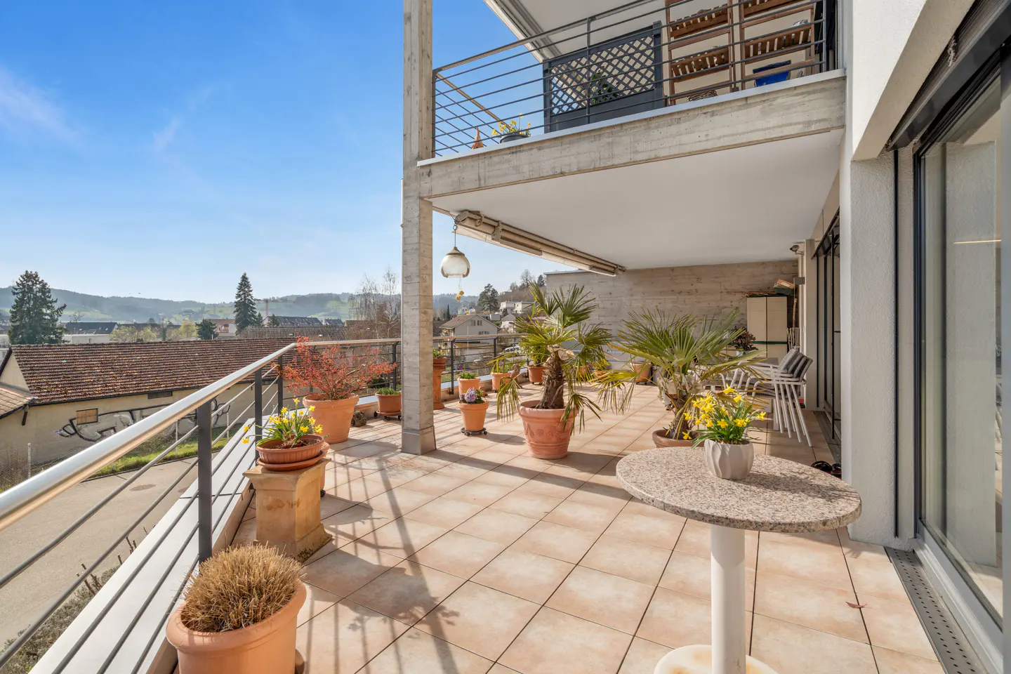 A sunny balcony with potted plants, a small table, and a view of a town.
