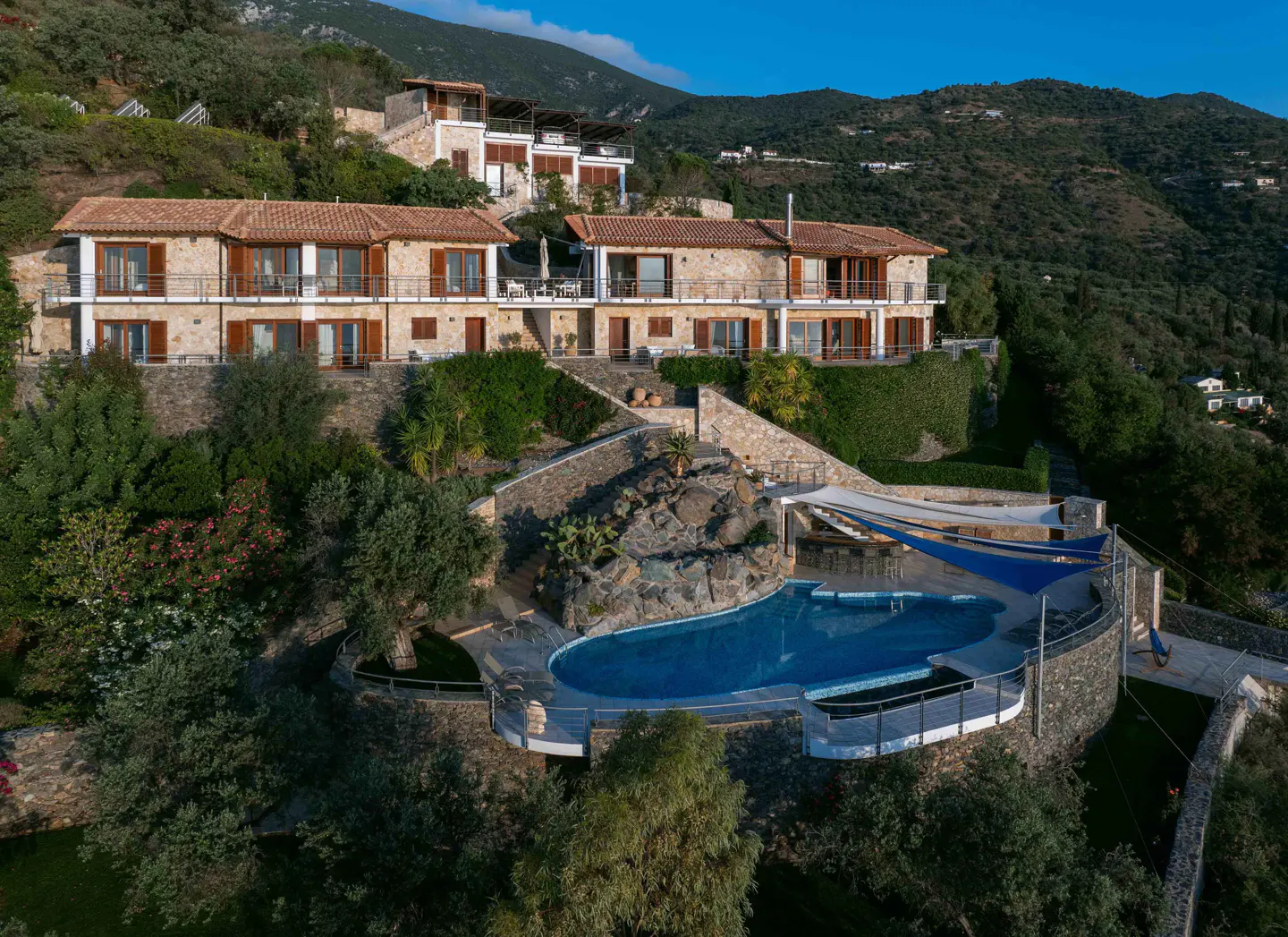 Luxury hillside villa with a pool. Stone buildings with red tile roofs are surrounded by lush green trees and mountains.