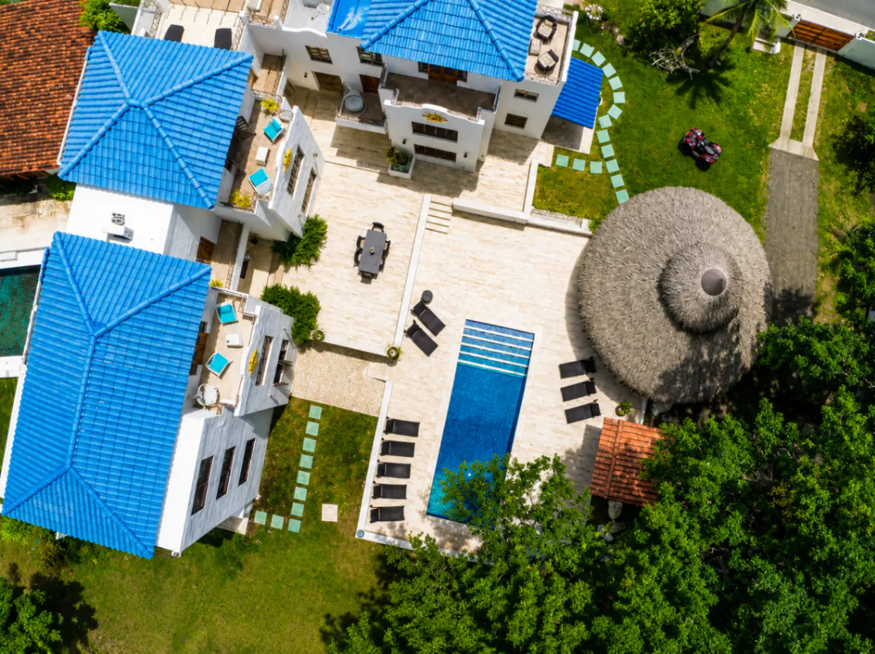Aerial view of a white villa with blue roofs, a pool, a thatched-roof gazebo, and green lawns.