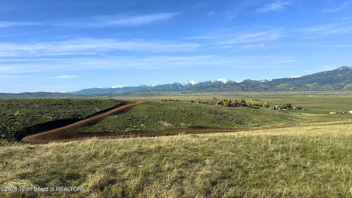 Scenic view of a grassy hillside with a dirt road, a valley with buildings, and snow-capped mountains under a blue sky.
