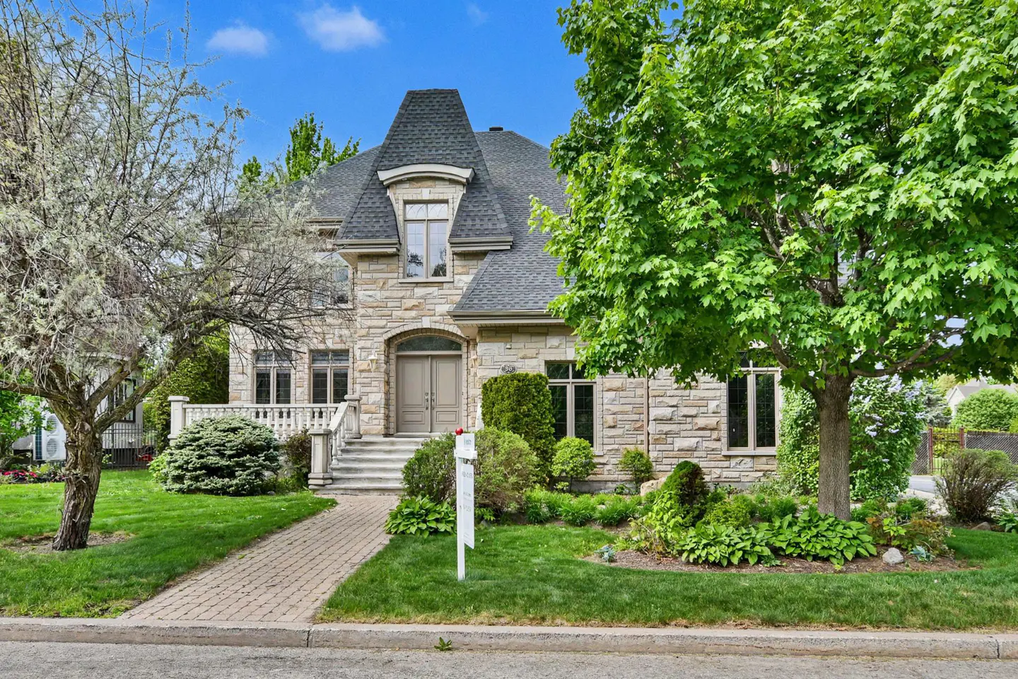 Two-story stone house with a gray roof, green lawn, and trees under a blue sky. A brick walkway leads to the front door.