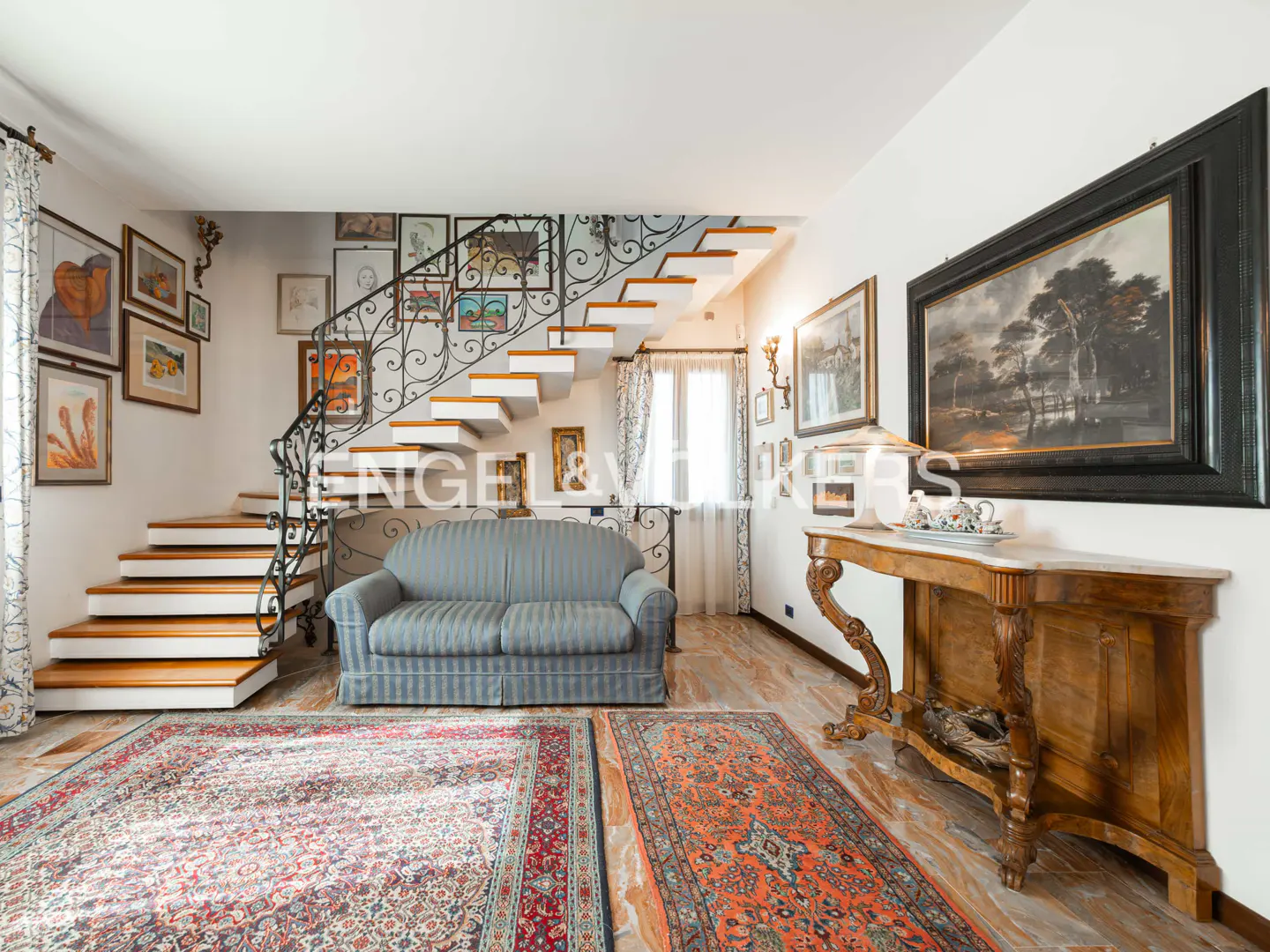 Living room with wooden stairs, blue sofa, and ornate black iron railing. Walls are decorated with framed art. Two rugs cover the marble floor.