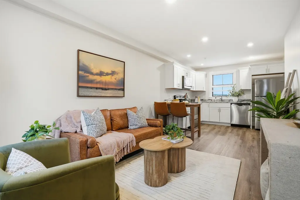 Open-concept living space with a brown leather sofa, green armchair, and white kitchen. A sunset painting hangs on the wall.