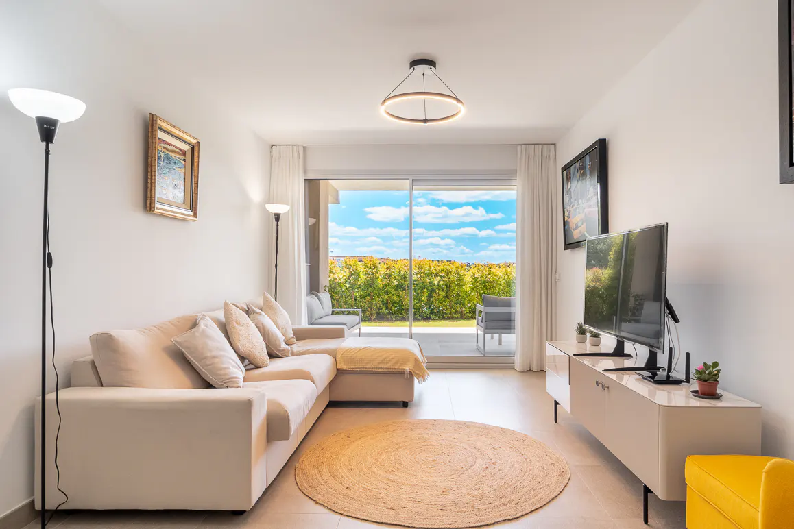 Bright living room with a beige sectional sofa, round jute rug, and a TV on a white cabinet. Sliding glass doors open to a patio with green hedges.