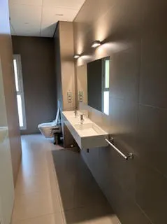 Modern bathroom with gray tile walls, a long white double sink, and a toilet in the background.