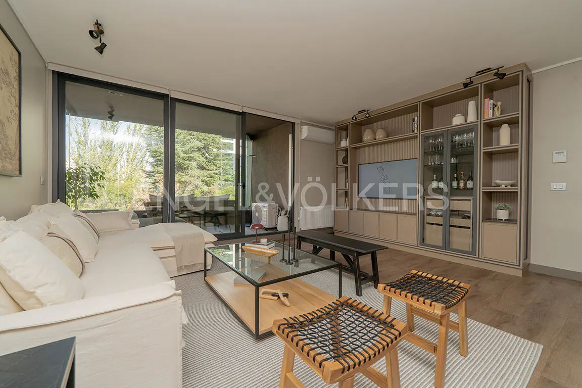 Living room with a white sofa, glass table, and wooden stools. A large window overlooks greenery. Built-in shelving unit with a TV and bar area.