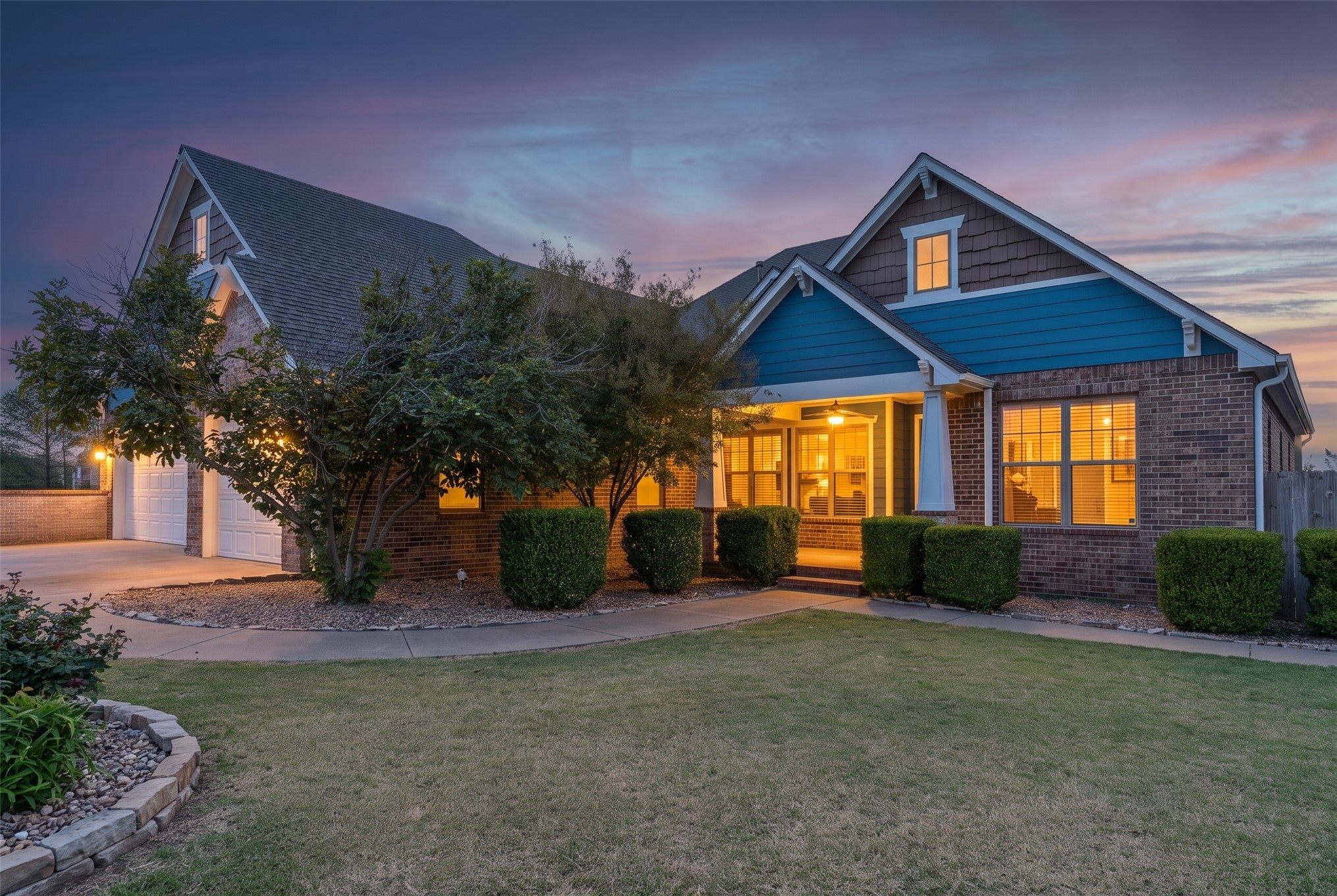 Brick house with blue siding and a lit porch at dusk, featuring a manicured lawn, trimmed hedges, and a two-car garage. [AI-generated text]