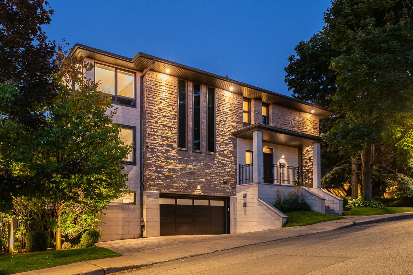 A two-story stone house with a black garage door and a small porch at dusk.