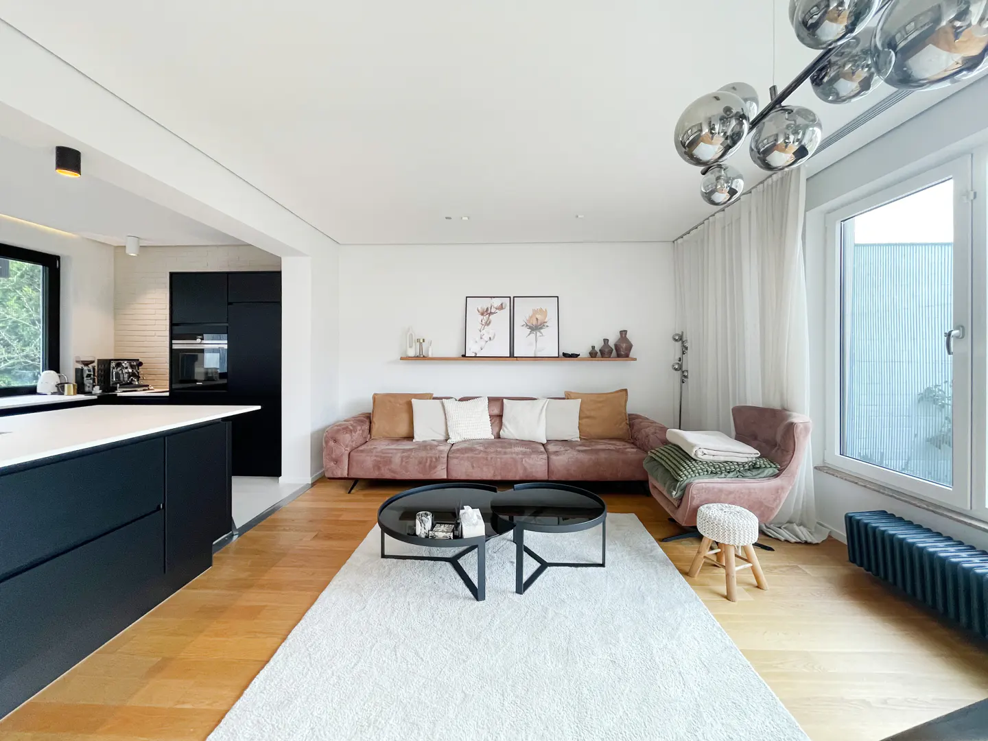 Open-concept living room with hardwood floors, white walls, and a pink sofa. Black kitchen island and modern lighting fixture.