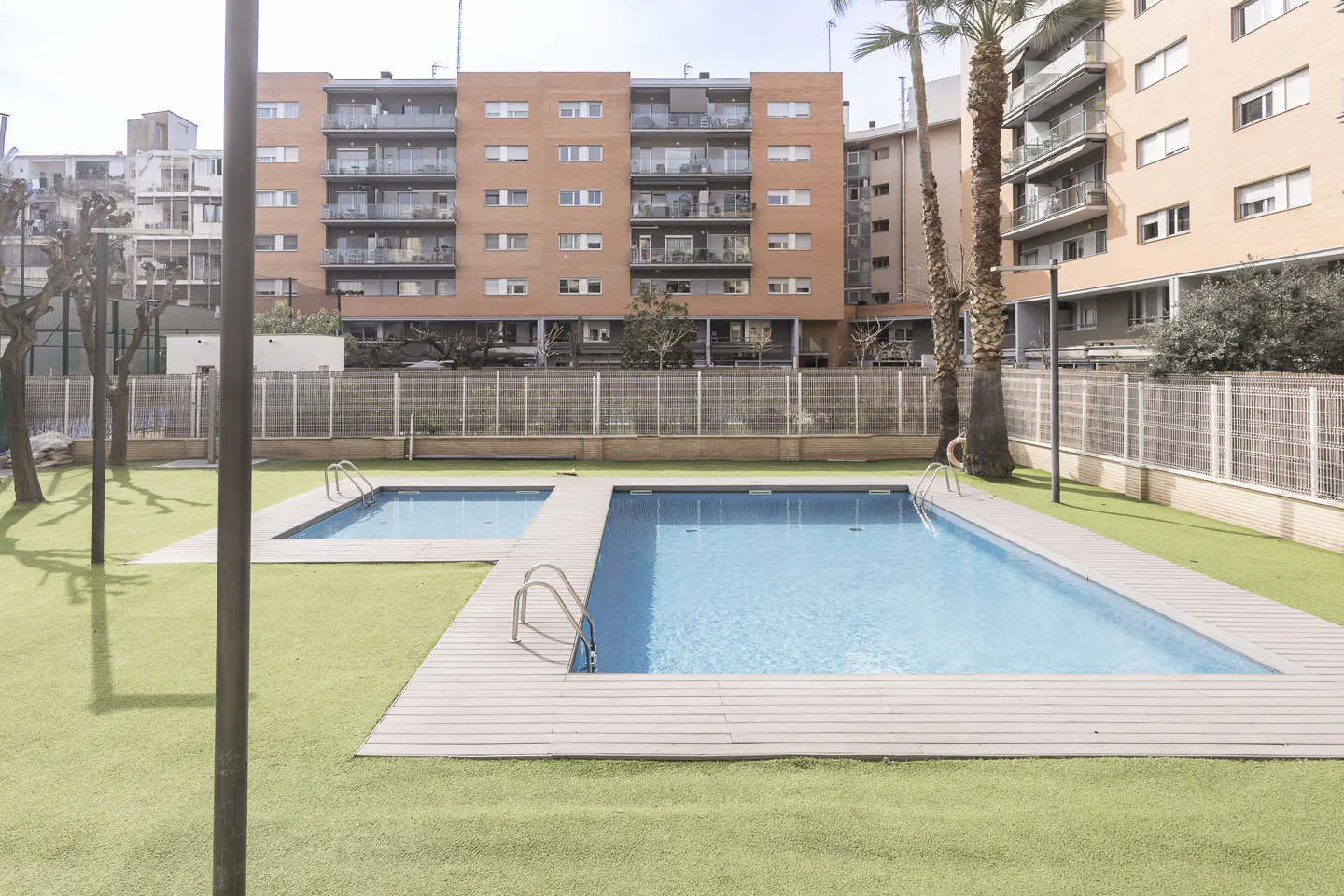 Outdoor pools with blue water and metal ladders sit on green turf, surrounded by a fence and apartment buildings.