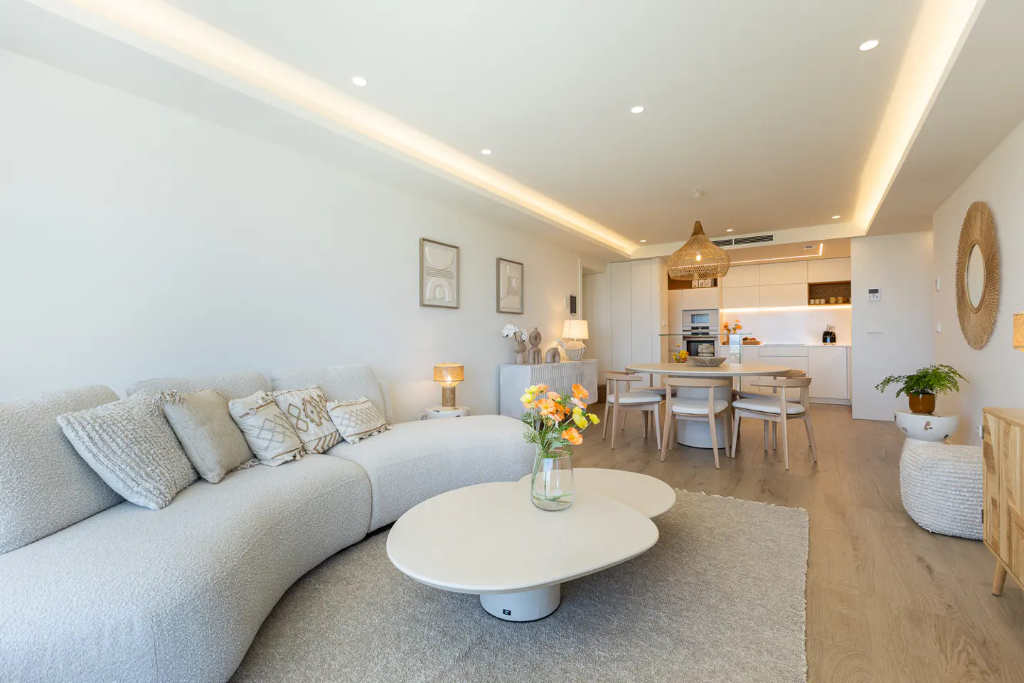 Bright, open-concept living space with a curved white sofa, modern coffee table, and dining area with a woven pendant light.