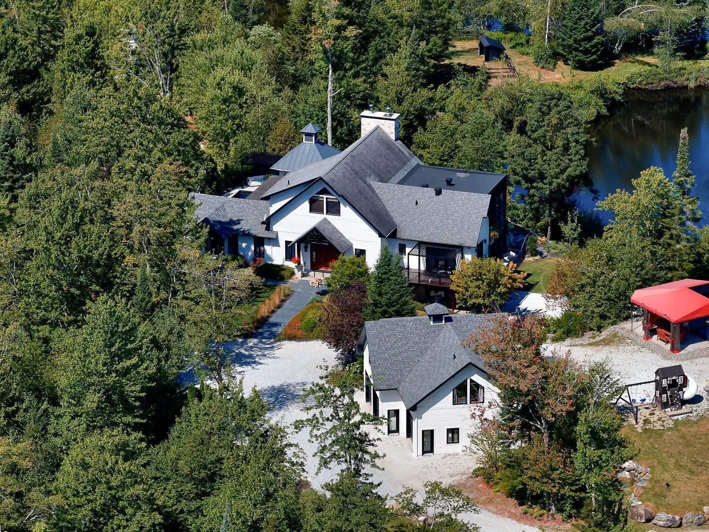 Aerial view of a large white house with a gray roof surrounded by green trees and a lake.