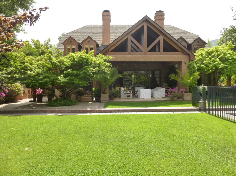 Backyard view of a brick house with a large lawn, patio, and trees. The patio furniture is covered.