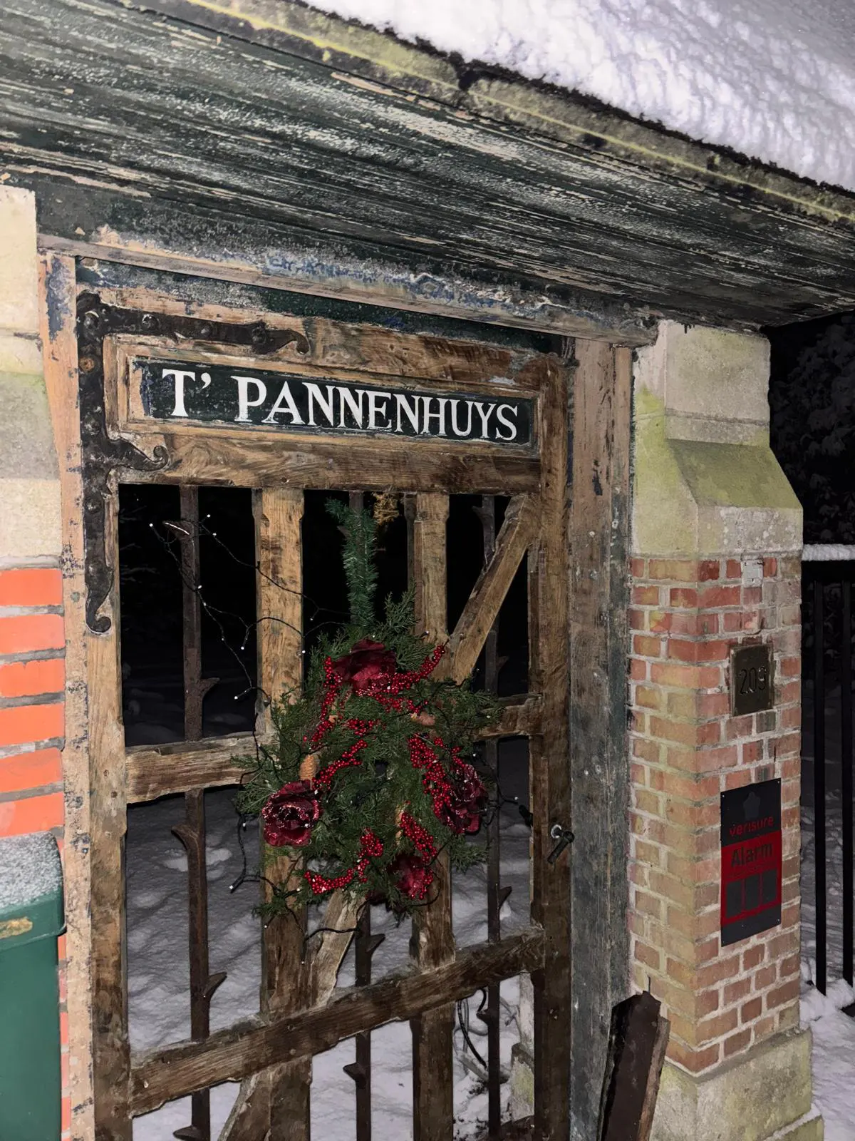 A wooden gate with a sign reading "T' PANNENHUYS" and a festive red and green wreath, set in a snowy brick entrance.