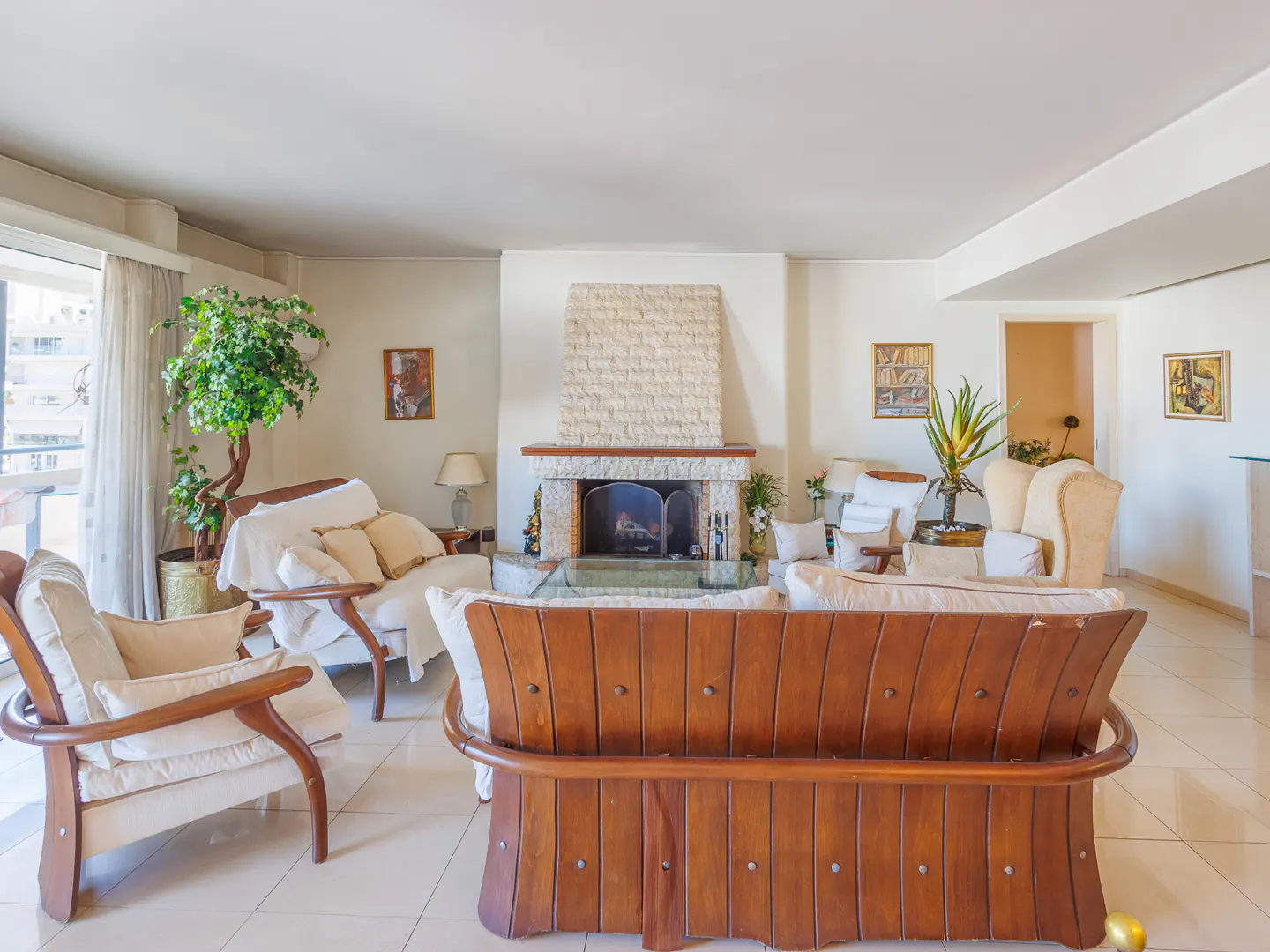 Bright living room with white walls, tile floors, and a stone fireplace. Wood-framed sofas and chairs with white cushions are arranged around a glass coffee table. A large window offers a view of the city.