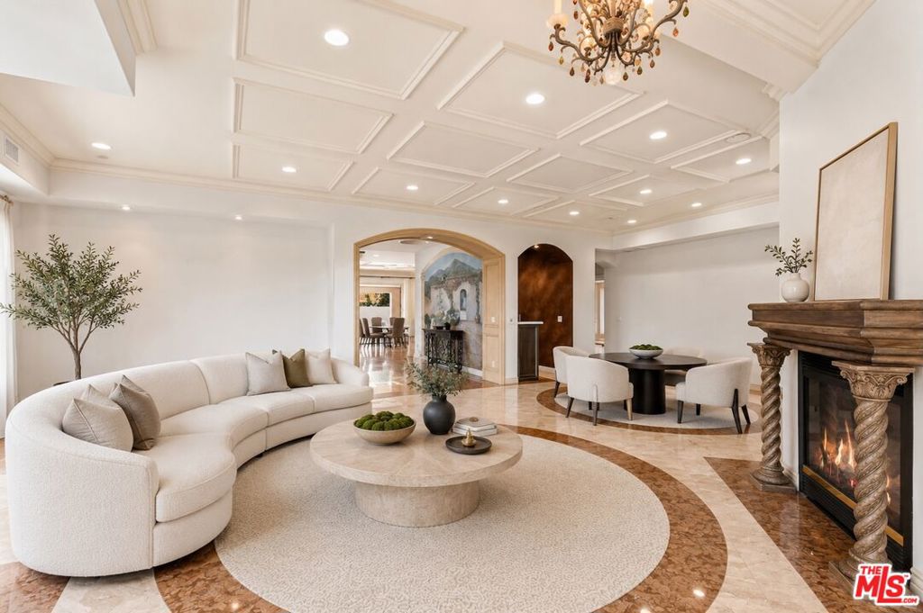 Elegant living room with a curved white sofa, round stone coffee table, fireplace, and dining area under a coffered ceiling.