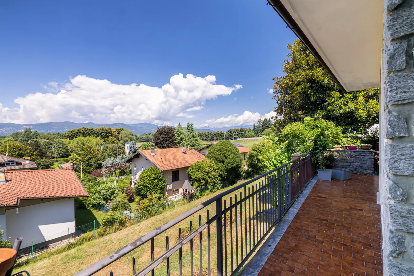 View from a balcony with a metal railing, overlooking houses, trees, and mountains under a blue sky with white clouds.
