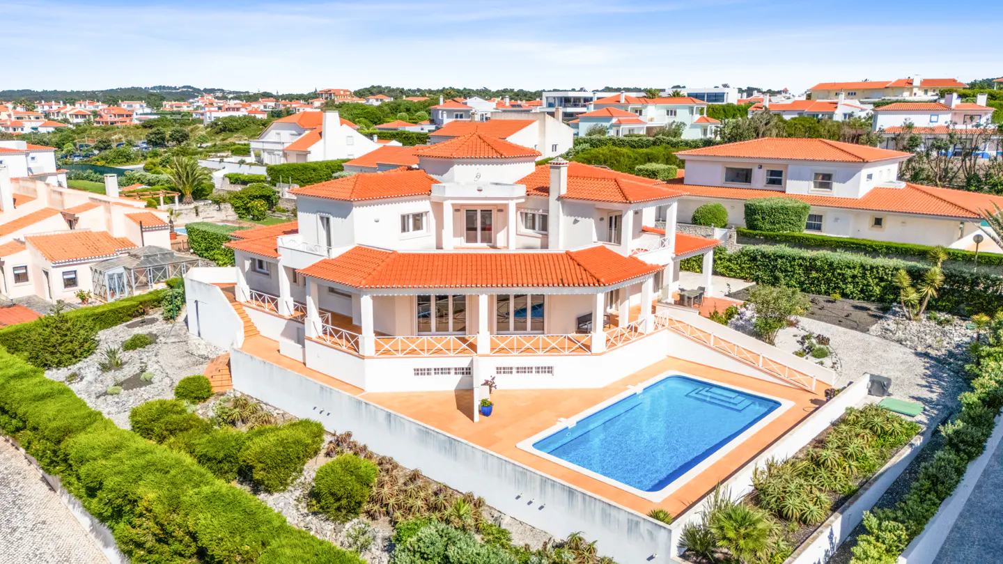 Aerial view of a white, multi-story house with an orange tile roof and a blue swimming pool in the backyard.