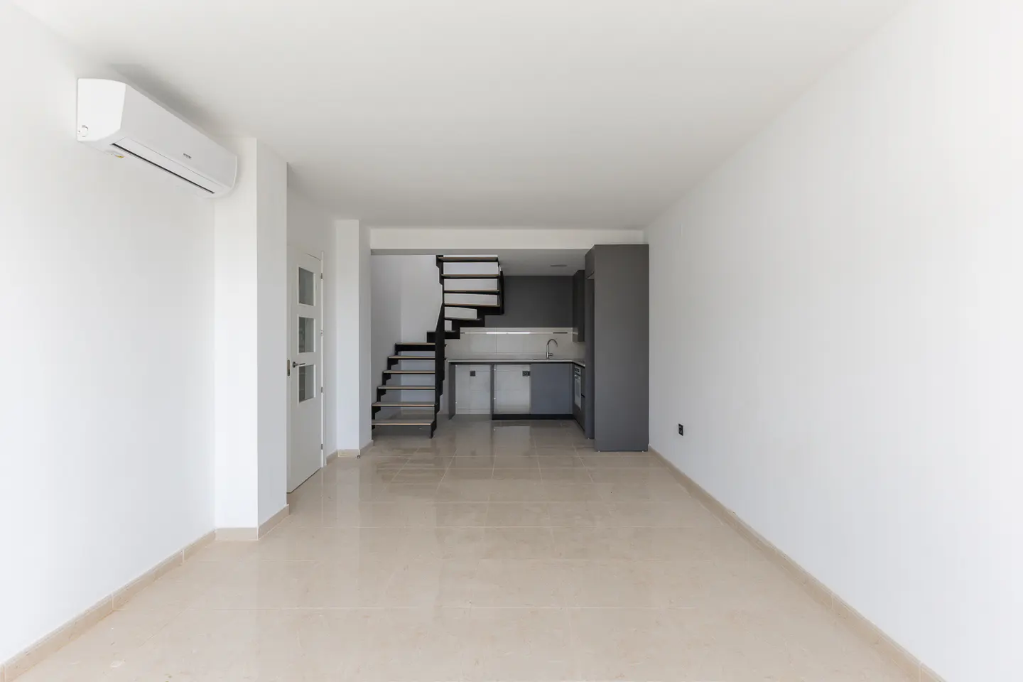 Bright, empty room with white walls, beige tile floor, and a modern gray kitchen and black spiral staircase in the background.