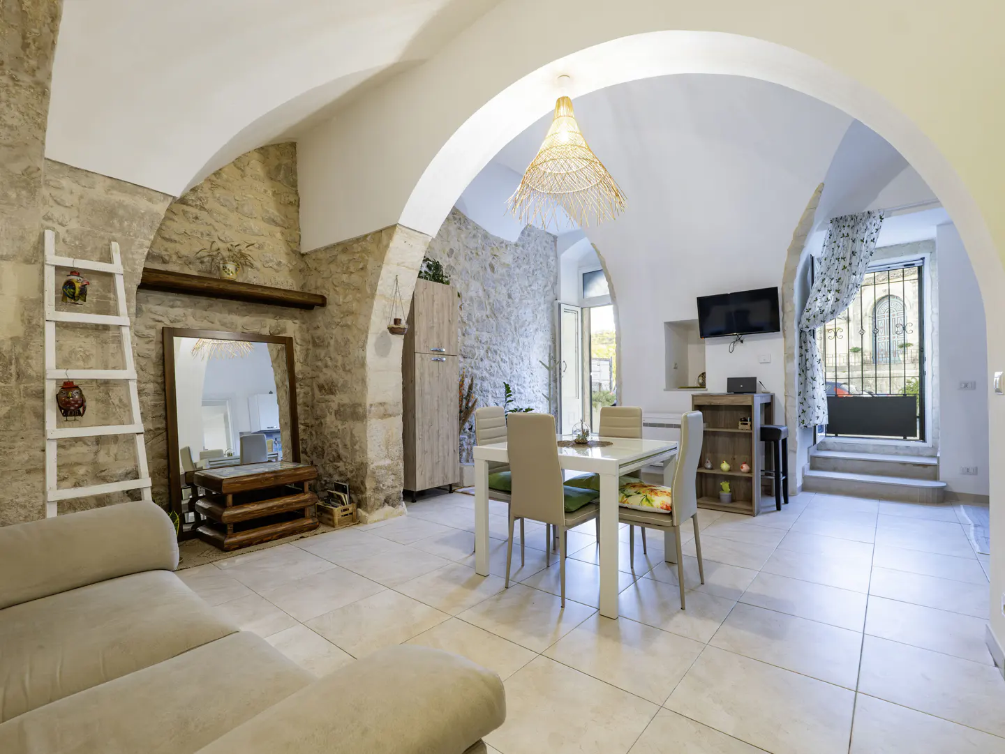 Bright living room with stone walls, arched ceilings, and tile floors. A white dining table sits near an open door with a wrought iron gate.