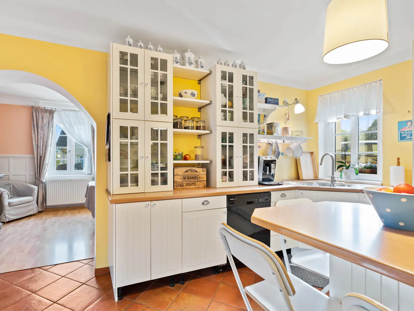 Bright kitchen with yellow walls, white cabinets, and terracotta tile floor. A doorway leads to a living room with a window.