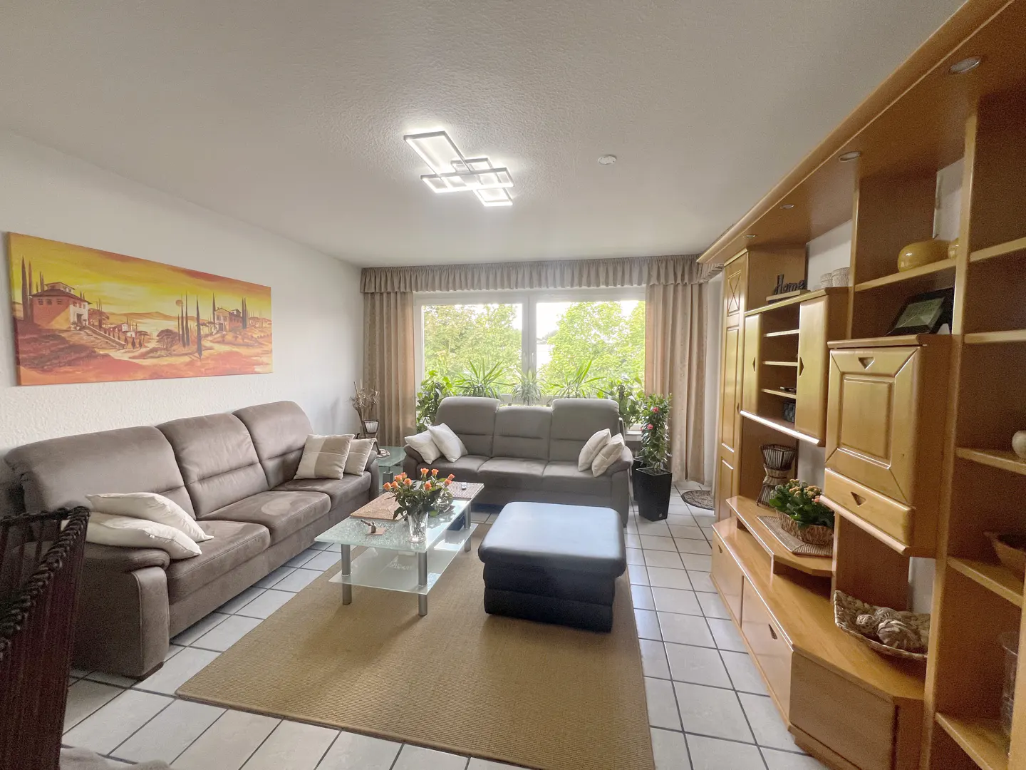 Living room with brown sofas, glass table, and wooden cabinet. A landscape painting hangs above the sofa. Large windows offer a view of green trees.