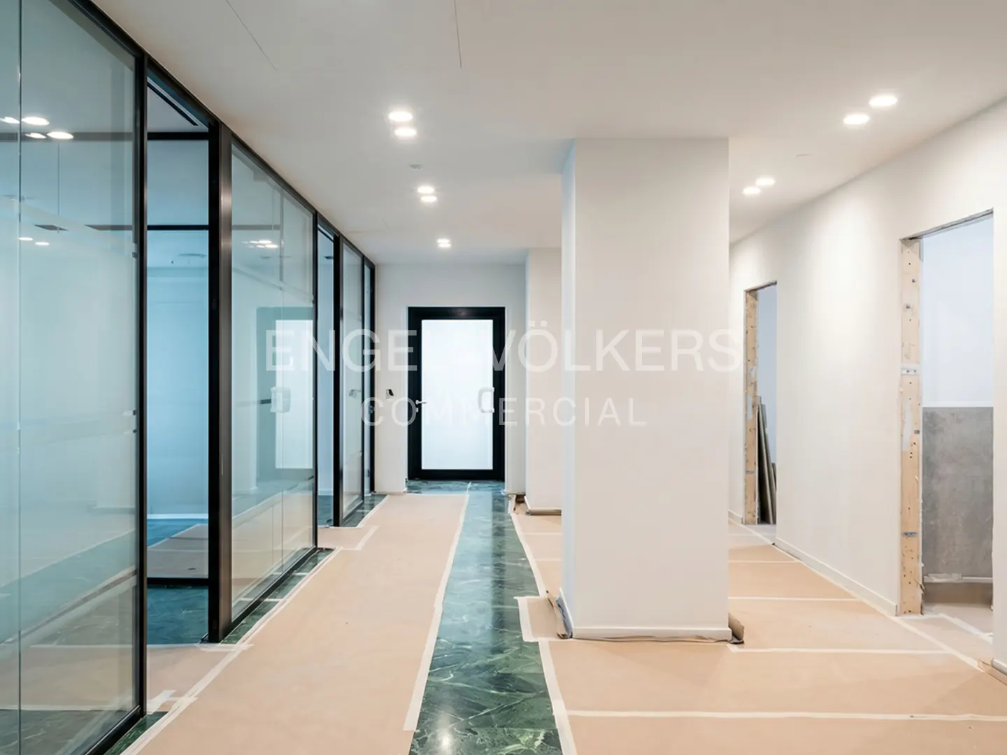 A bright office hallway with glass-walled offices on the left and a black door at the end. The floor is green marble.