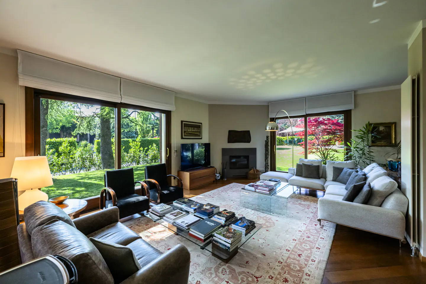 A bright living room with a brown leather sofa, black chairs, and a white sectional. Large windows overlook a green lawn and trees. A glass coffee table holds books.