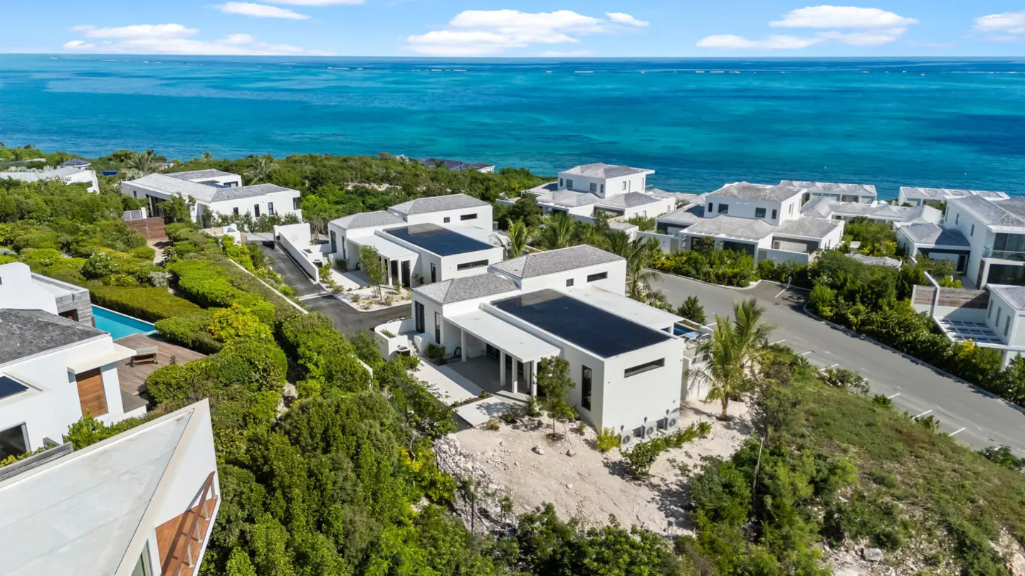 Aerial view of modern white villas with gray roofs, surrounded by lush green vegetation, overlooking the turquoise ocean under a blue sky.
