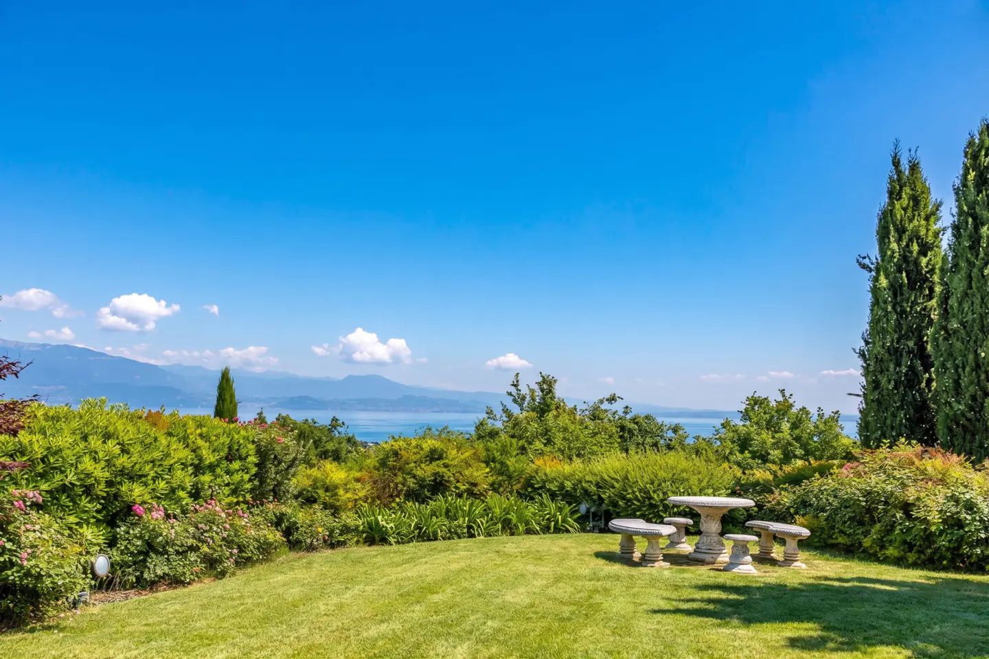 Outdoor patio with stone table and benches on green lawn, surrounded by lush greenery, with a lake and mountains in the background under a blue sky.