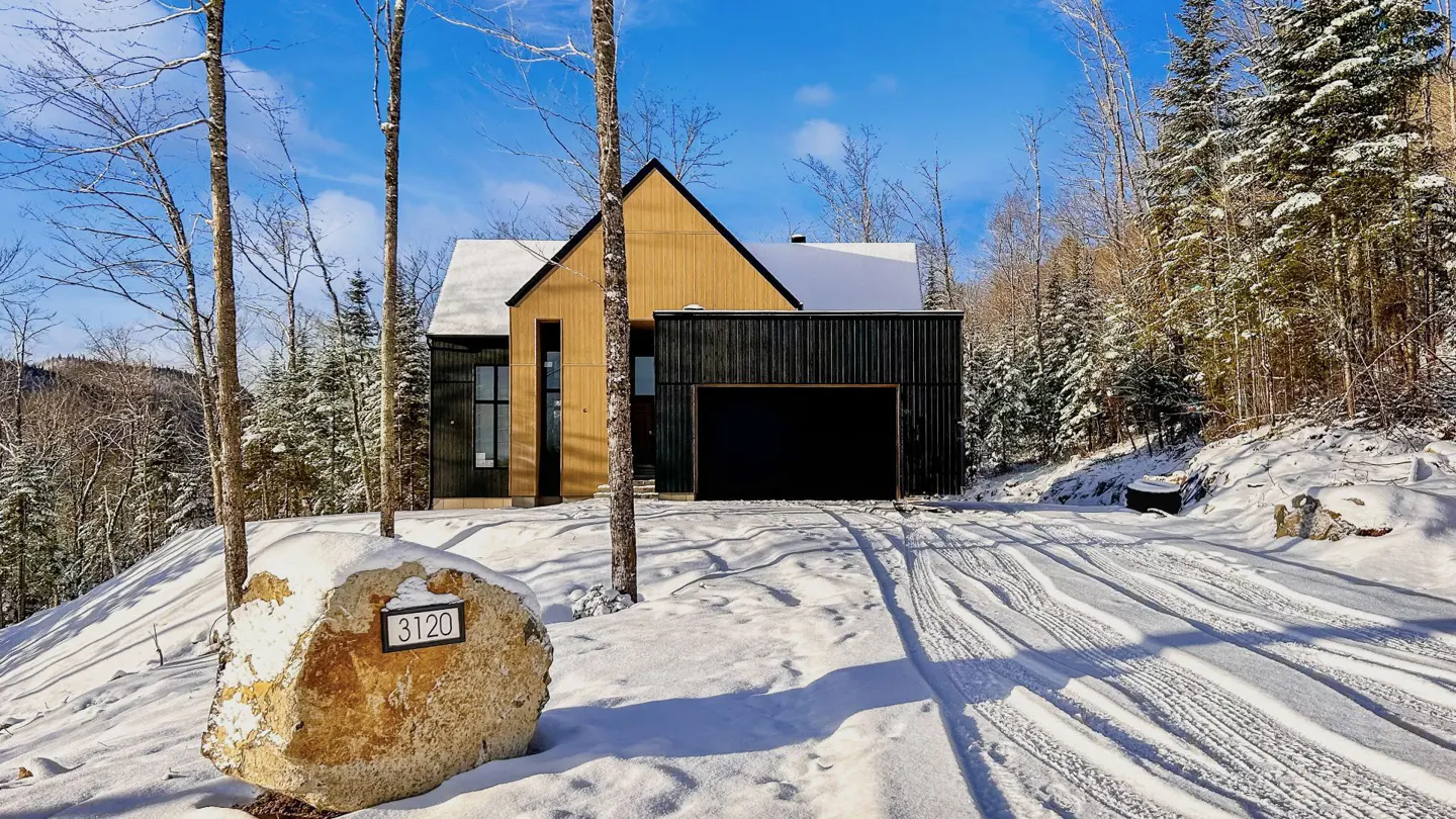 Modern home with light wood and black siding, snow-covered roof, and a black garage door. A stone with the number 3120 sits in the snowy yard.
