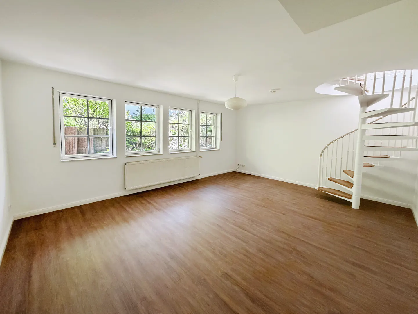 Bright, empty room with wood floors, white walls, and a spiral staircase. Three windows overlook a green yard.