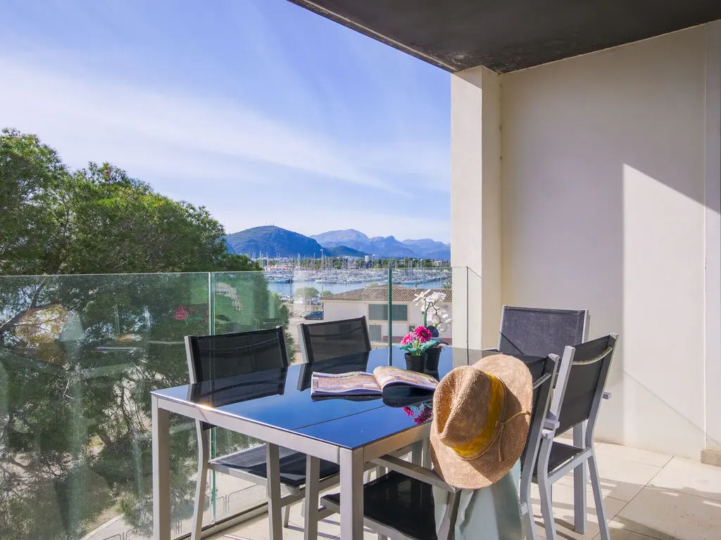 Balcony with a glass table, chairs, a hat, and a magazine overlooking a marina and mountains.