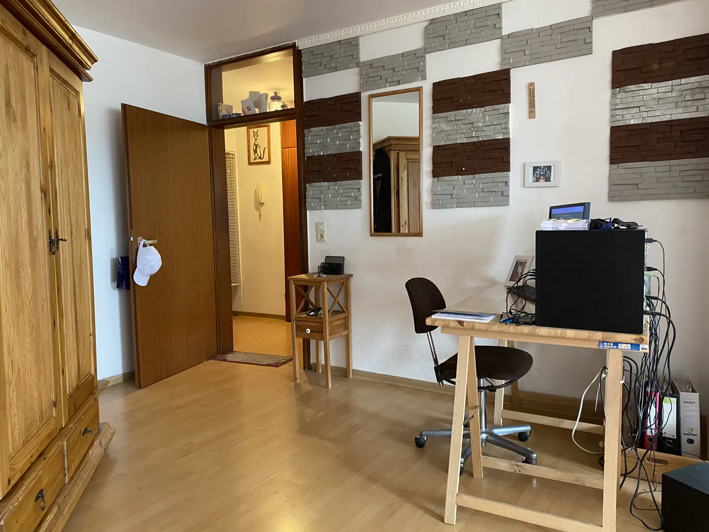 A room with a wooden floor, a desk with a computer, a chair, and a wooden wardrobe. The wall has decorative stone panels.