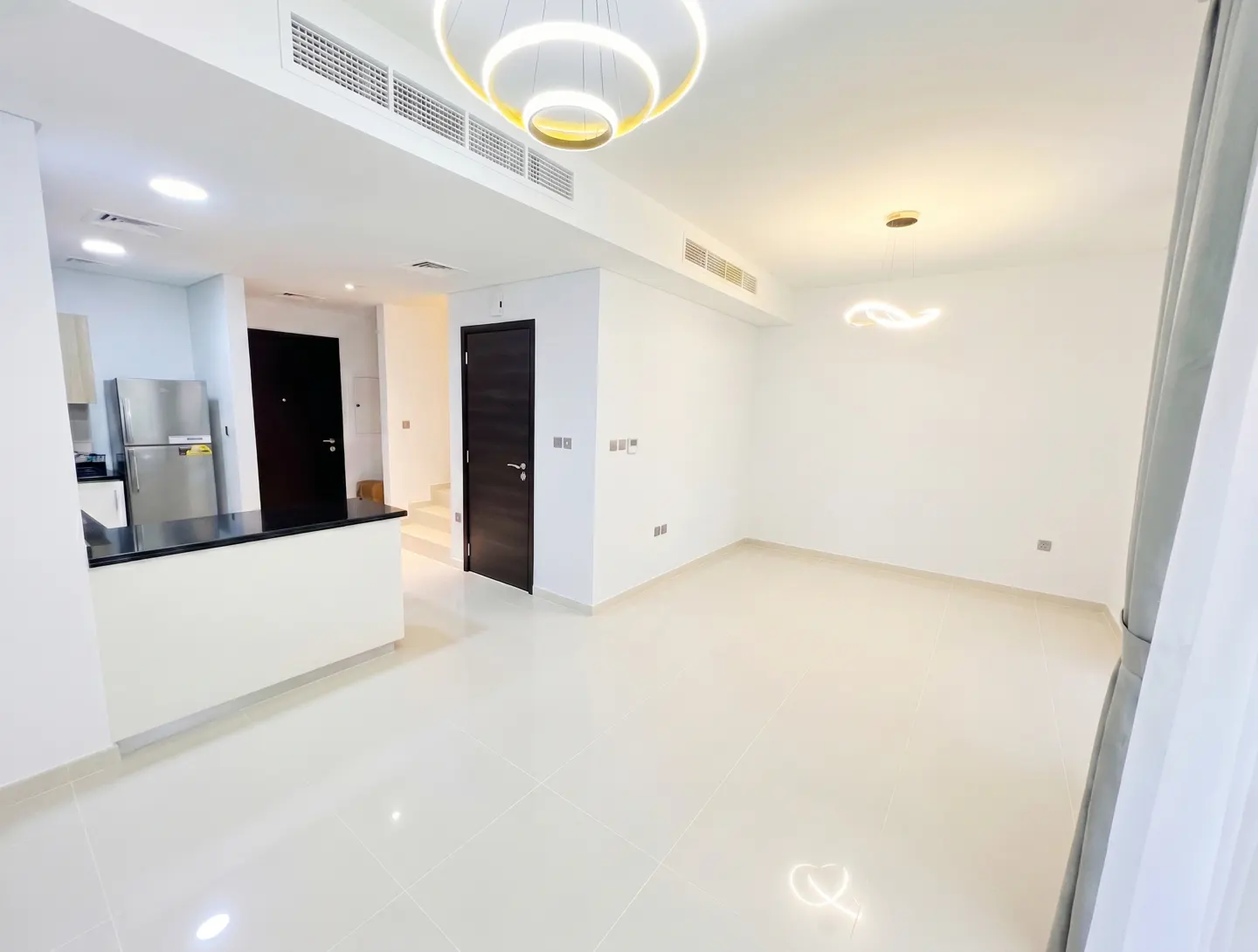Bright, modern apartment interior with white walls and tile floors. A kitchen area with stainless steel appliances and a black countertop is visible.