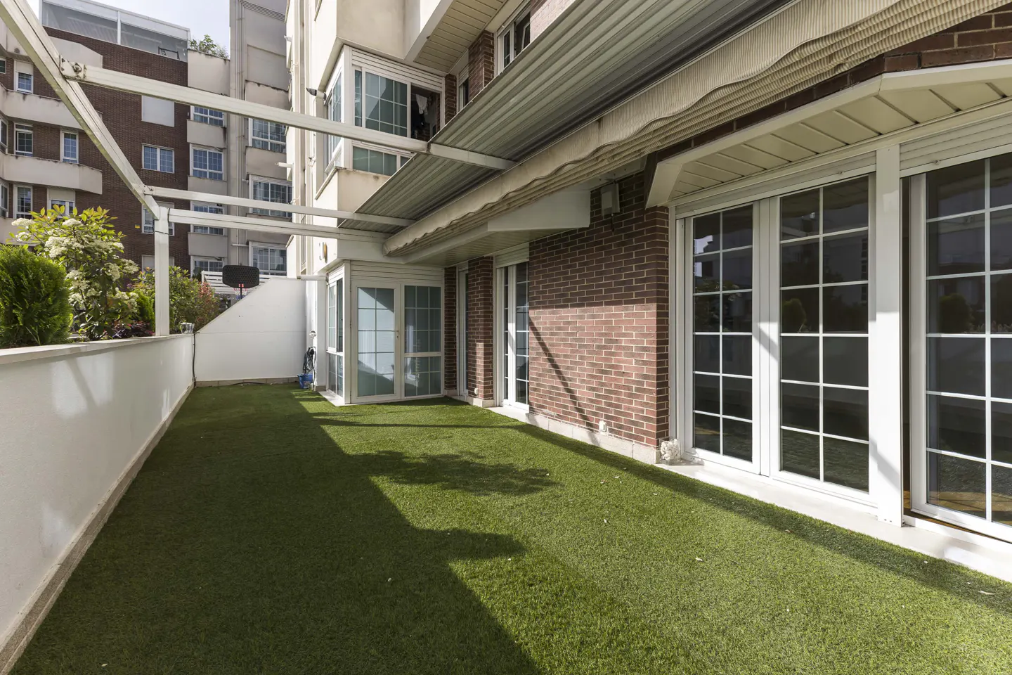 A patio with artificial grass, a brick wall, and white-framed glass doors. A white pergola covers part of the patio.
