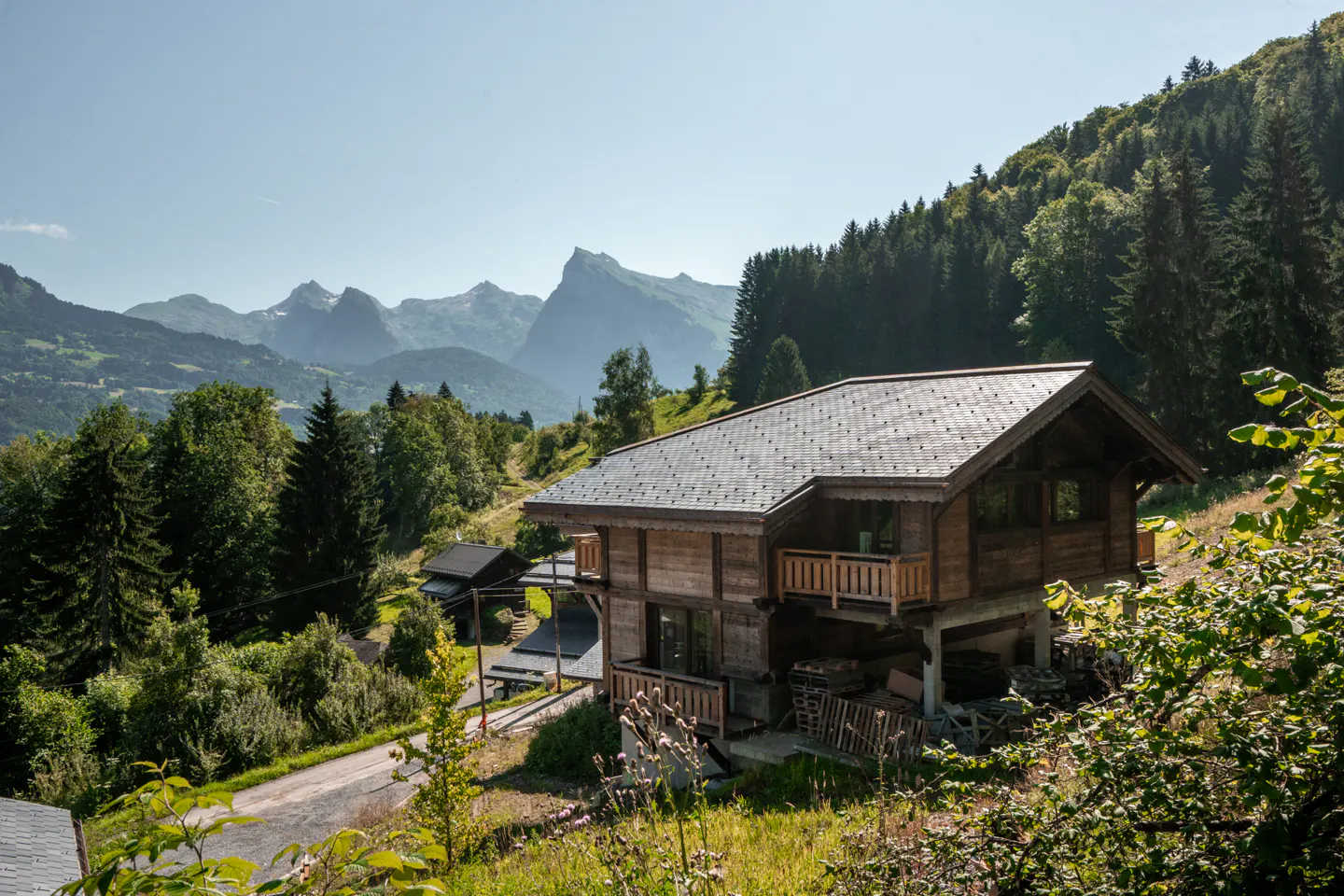 Exterior view of a two-story wood chalet with balconies, set against a backdrop of green trees and mountains.