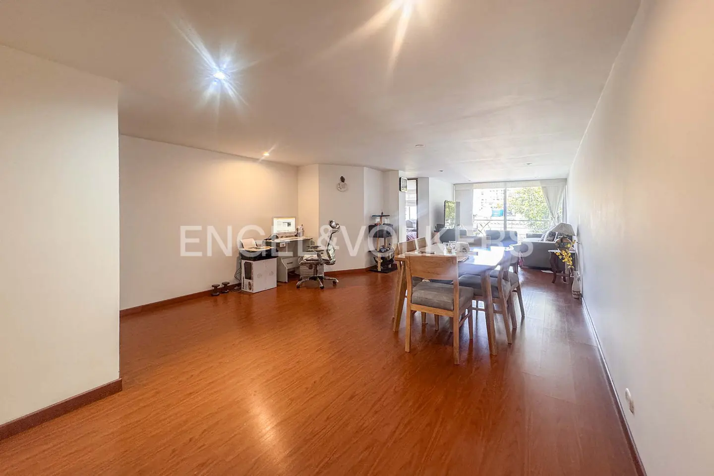 Open-concept living space with wood floors, white walls, dining table, and office area. Natural light fills the room.