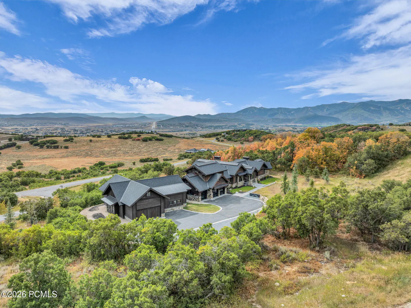 Aerial view of a dark brown house with a gray roof, surrounded by green trees, with mountains and a blue sky in the background.